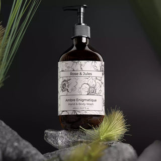 Flacon ambré avec pompe noire du gel douche bio et vegan "Ambre Enigmatique" de Rose et Jules, posé sur un rocher entouré d'herbes et de plantes, évoquant la nature.