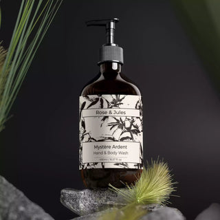 Flacon ambré avec pompe noire du gel douche bio et vegan "Mystère Ardent" de Rose et Jules, posé sur un rocher avec des herbes et plantes en arrière-plan, évoquant la nature.